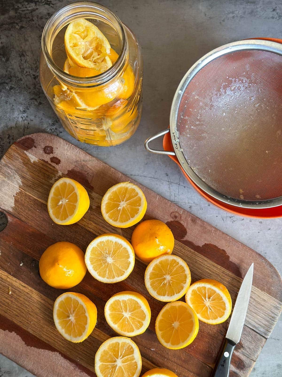 Citrus that has been cut in half is arranged on a wood cutting board with a knife. Above the board is a large mason jar filled with citrus peels along with a bowl and strainer.