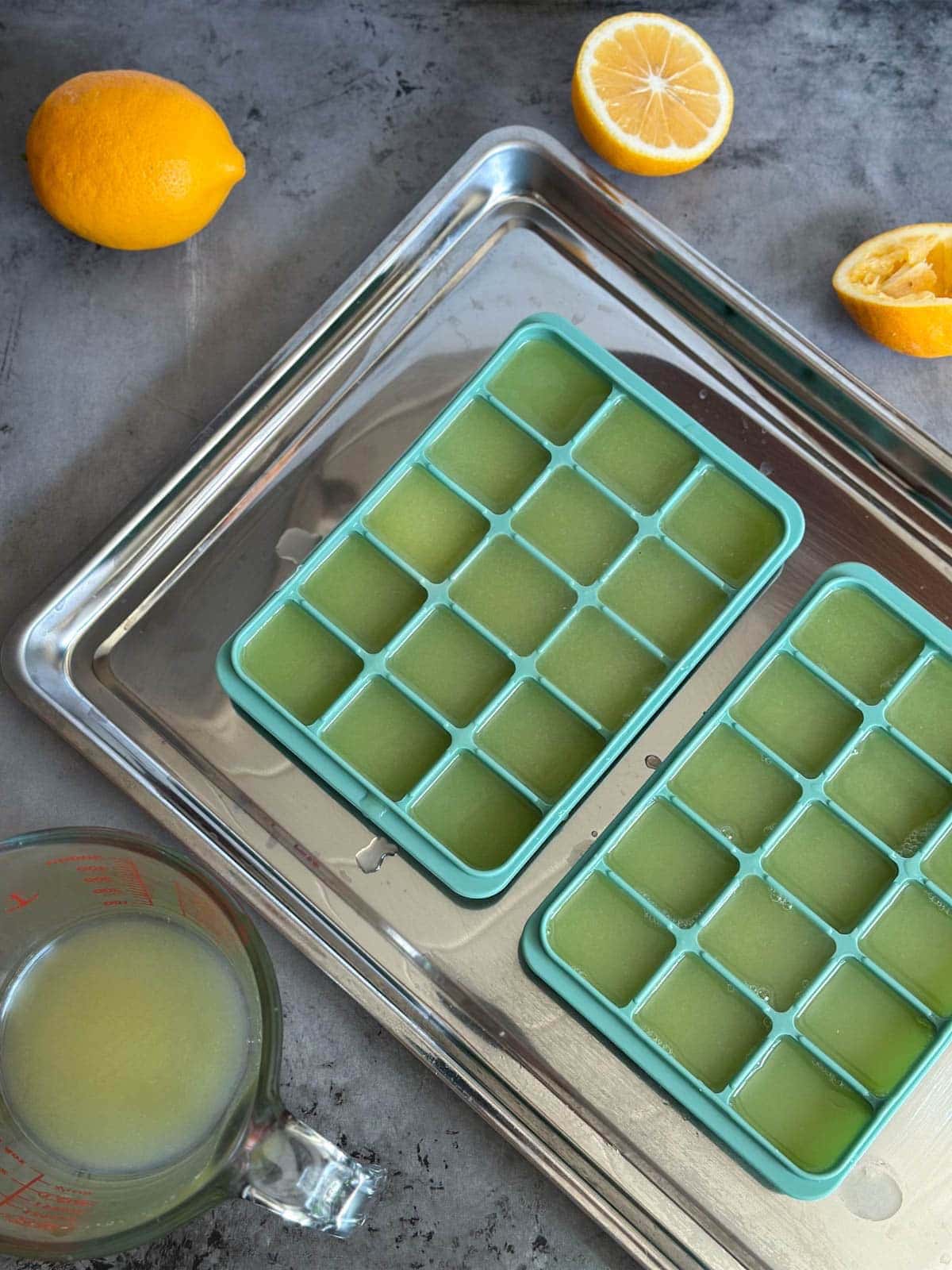 Two silicone ice cube trays filled with juice are sitting on a metal baking sheet. A liquid glass measuring cup along with a one whole lemon and one that has been cut in half surround the metal sheet.