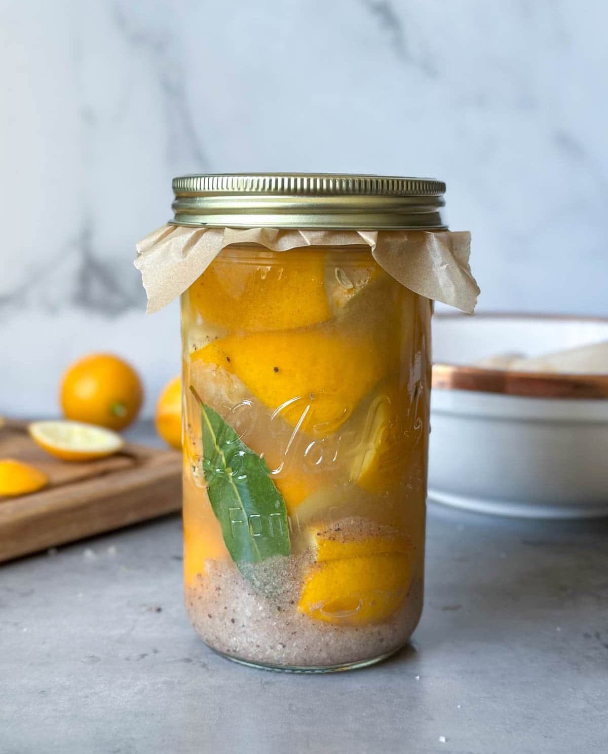 A one liter jar packed full of preserved lemons with a bay leaf inside sits in front of a wood cutting board with whole and sliced lemons.