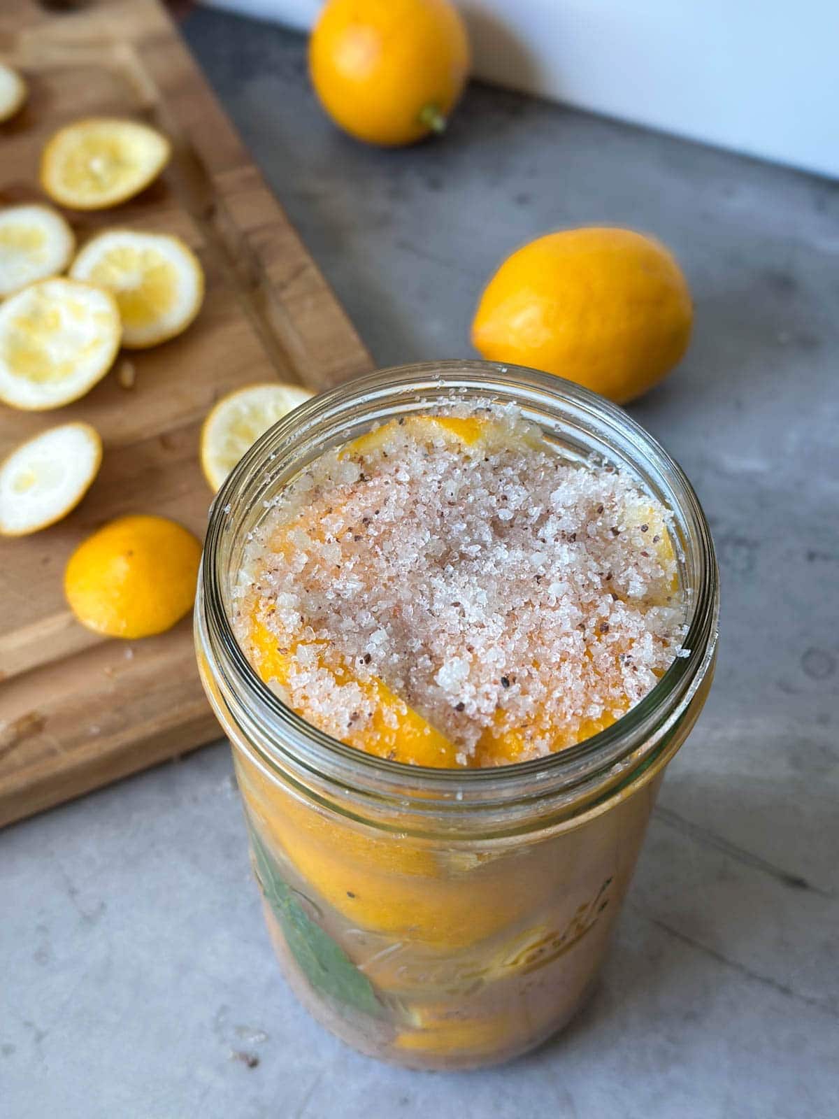 A layer of salt sits on top of citrus packed into a glass jar. Beyond is a wood cutting board with whole citrus and slices scattered about.