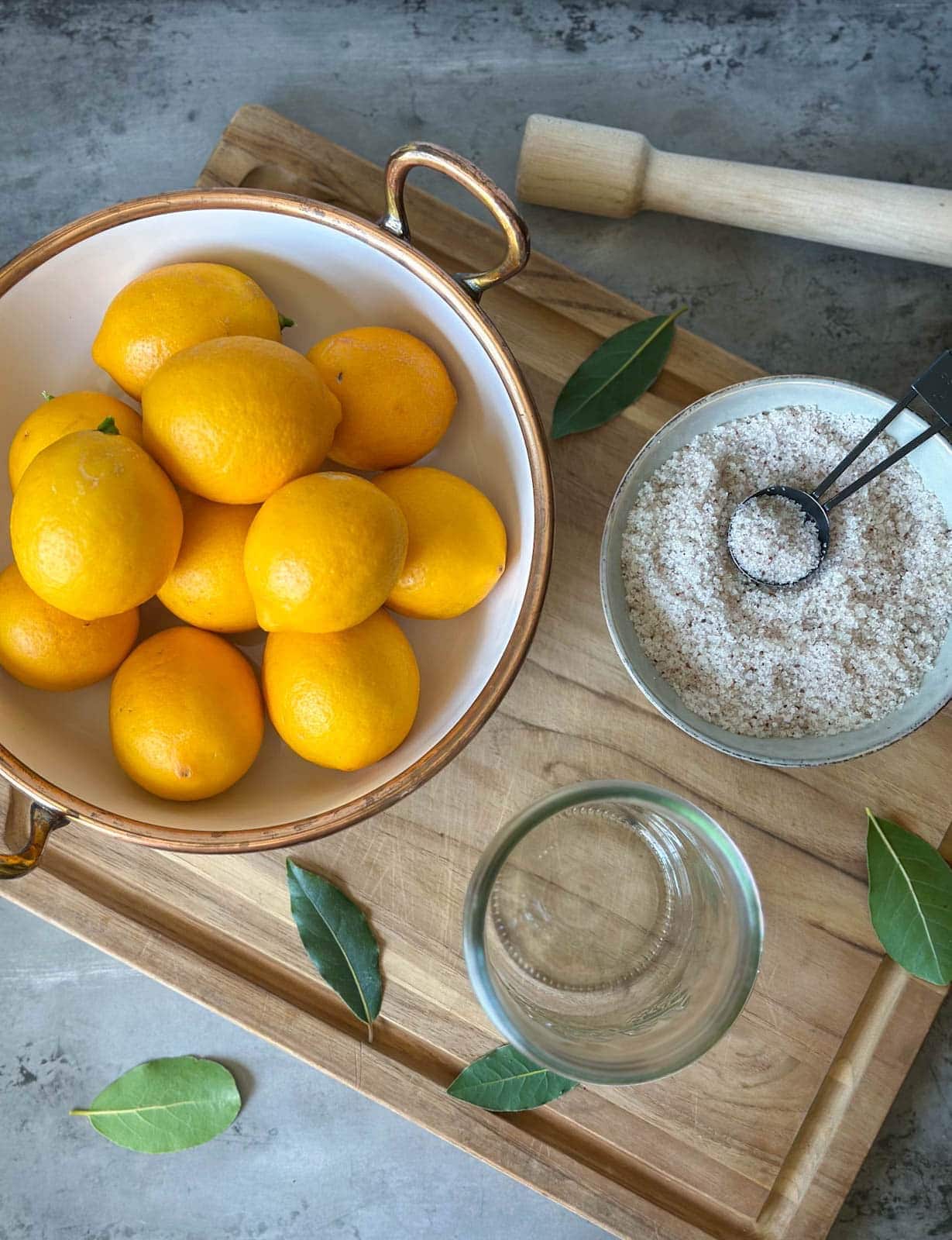 A bowl of Meyer lemons, a small bowl of salt, an empty glass jar, and a few sprigs of bay leaves are all arranged on a wood cutting board. A wood pounder sits just above the cutting board.