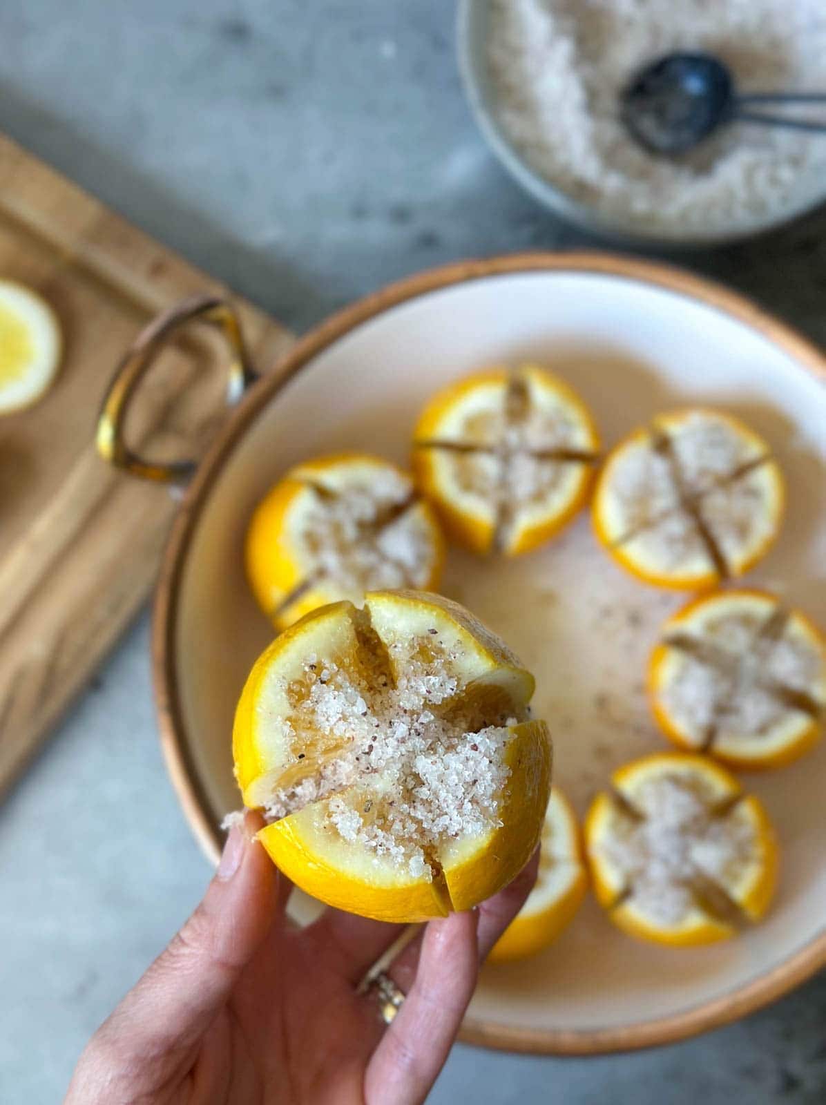 A hand is holding a whole lemon that has been cut into quarters while not cutting through all the way to leave the fruit in one piece. Salt is packed on top of and inside the segments. Below is a large bowl which containers more citrus that has been packed with salt.