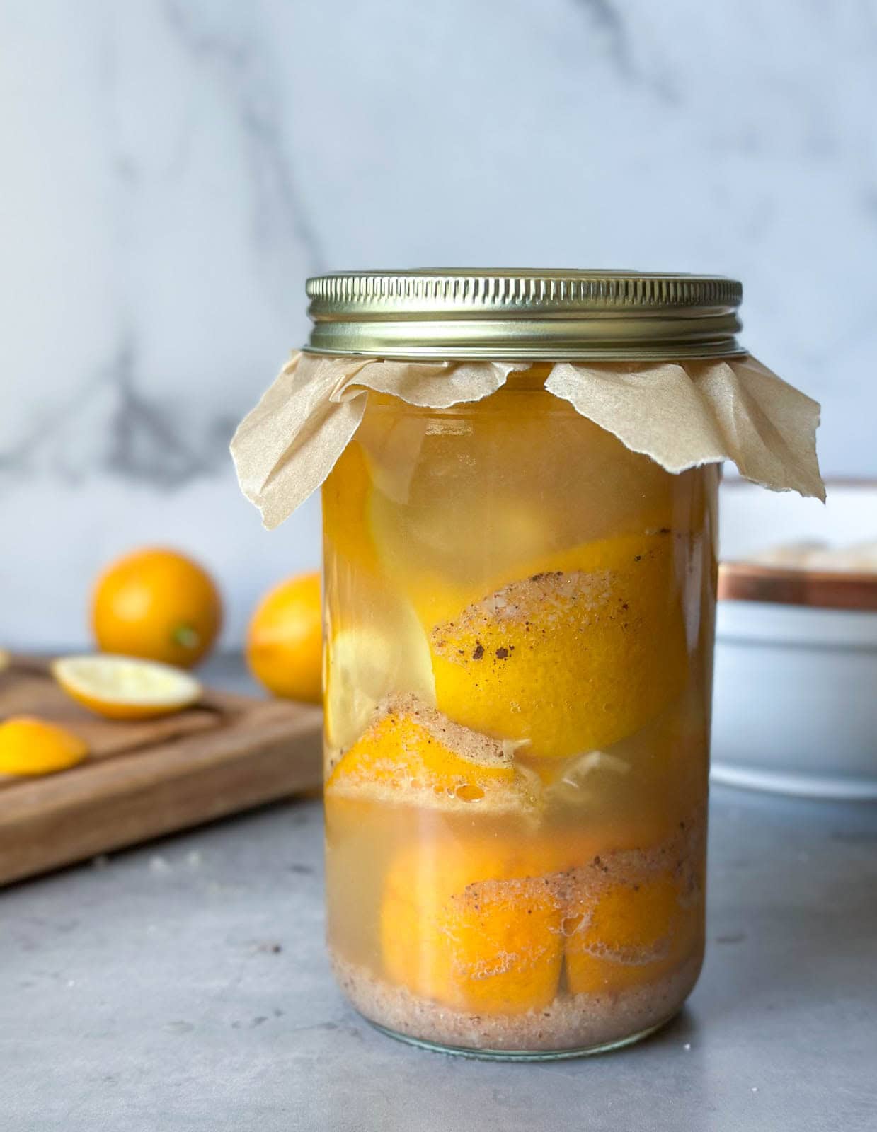 Preserved lemons packed with salt and lemon juice inside a one liter sized jar with a metal lid with a piece of parchment paper underneath. Beyond is a wood cutting board with sliced lemons and a few whole lemons.