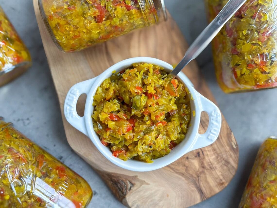 A white ramekin container is full of zucchini relish. A few canned jars of relish lay around the outside of the ramekin.