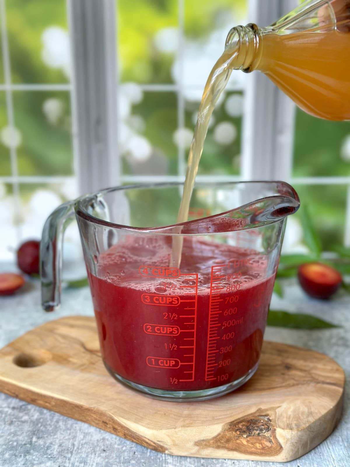 A bottle of apple cider vinegar is being poured into two cup glass liquid measuring cup that contains a plum syrup to make a shrub recipe. 