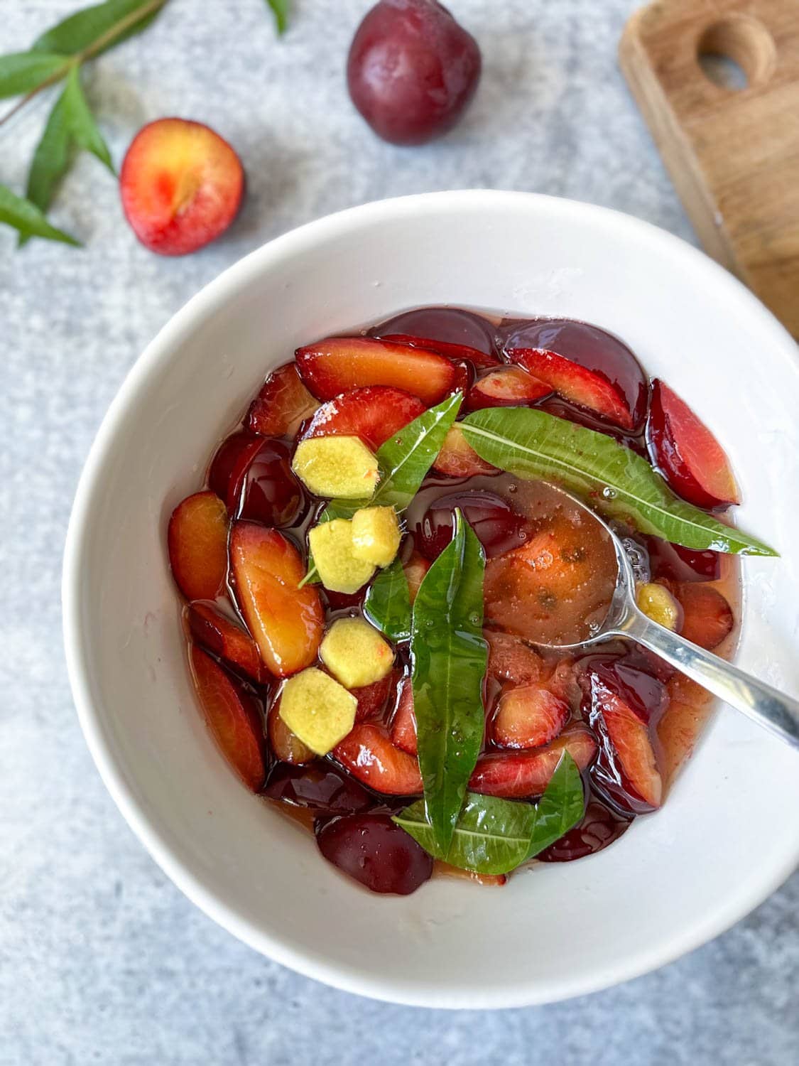 A white ceramic bowl contains sliced plums, lemon verbena, and chunks of fresh ginger. Lemon verbena and a few plums garnish the surrounding area. 
