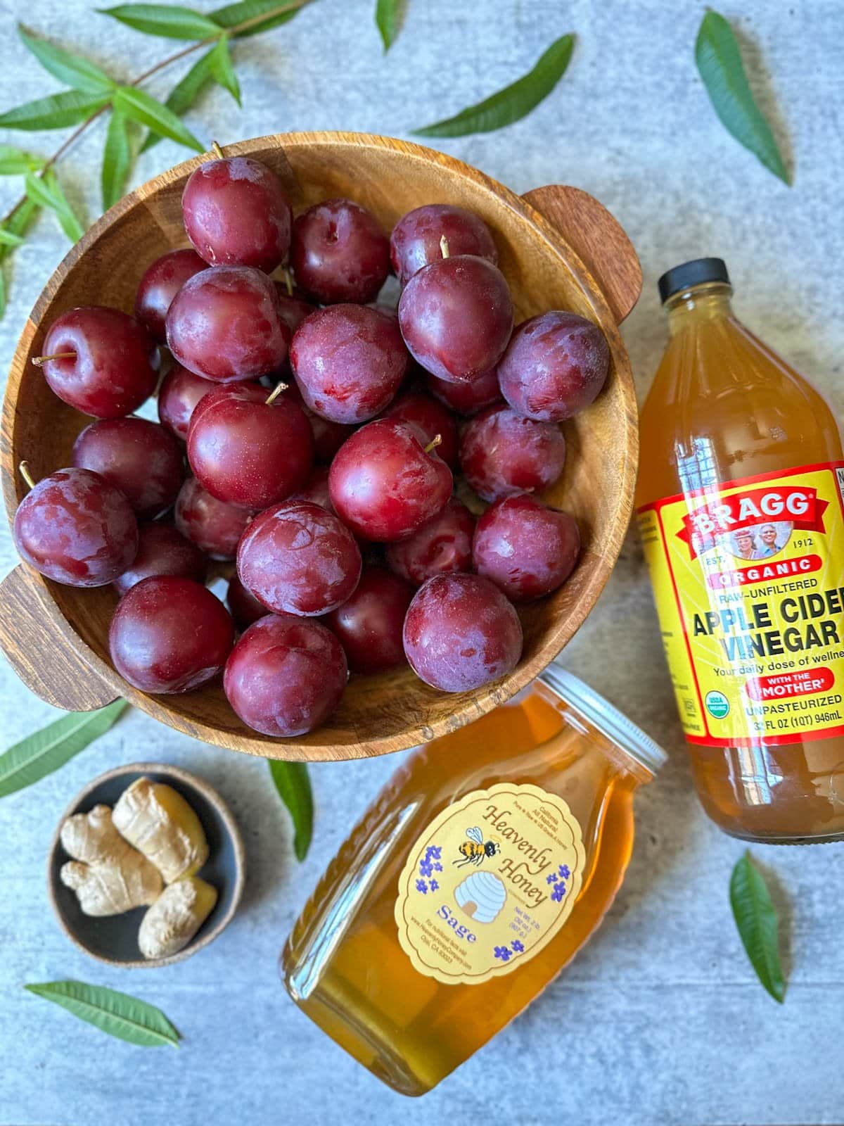 A bowl of harvested plums is surrounded by a bottle of apple cider vinegar, a jar of honey, chunks of fresh ginger, and lemon verbena leaves make up the contents of the fruit shrub recipe. 