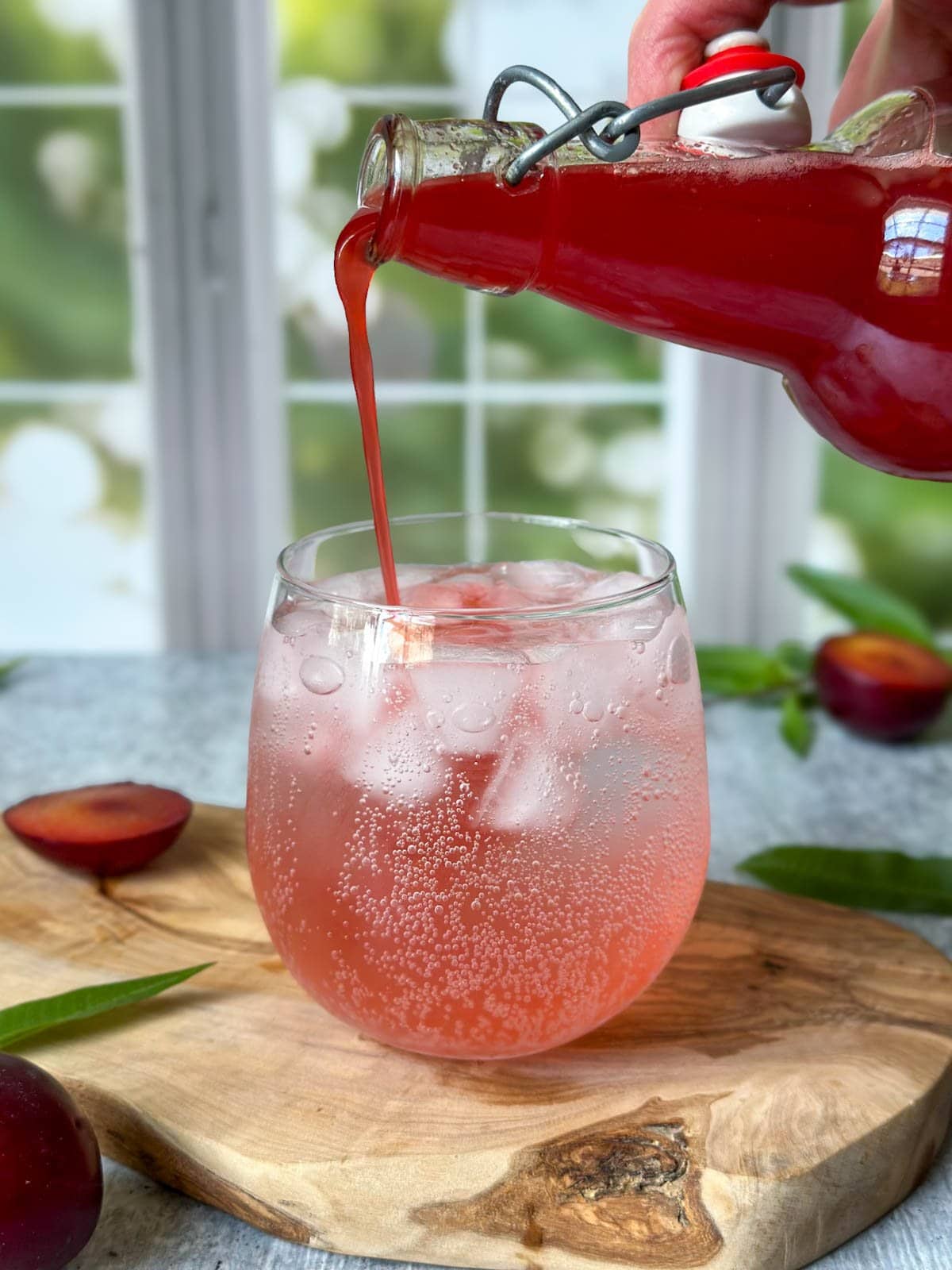 A flip top bottle filled with fruit shrub being poured into a stemless wine glass full of sparkling water. Plum and lemon verbena decorate the surrounding area. 