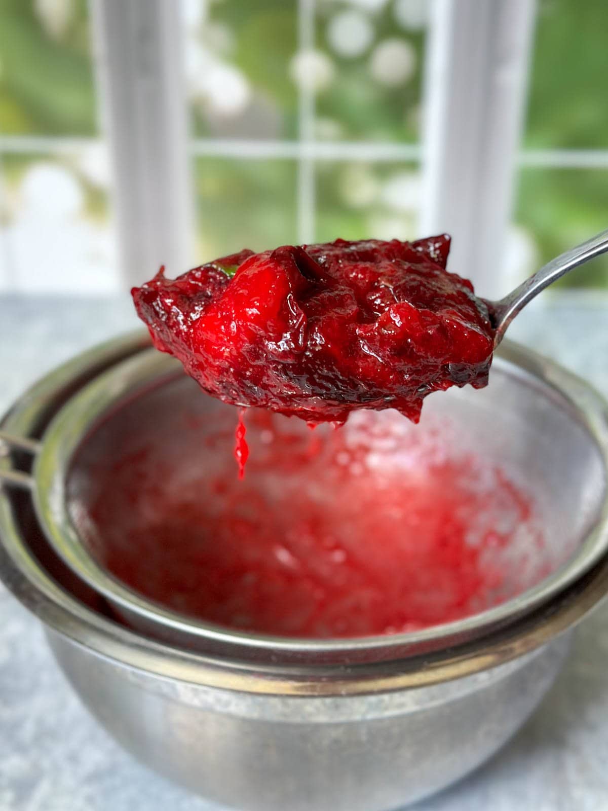A spoonful of plum skins and ginger chunks is held above a metal bowl with a fine mesh stainless steel strainer placed on top of it. 