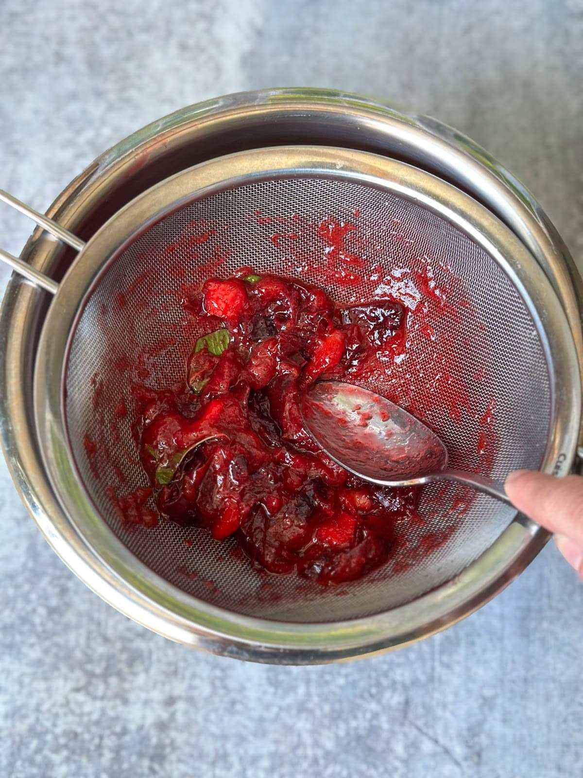 A large spoon is being used to press macerated fruit through a fine mesh strainer with a metal bowl beneath it. 