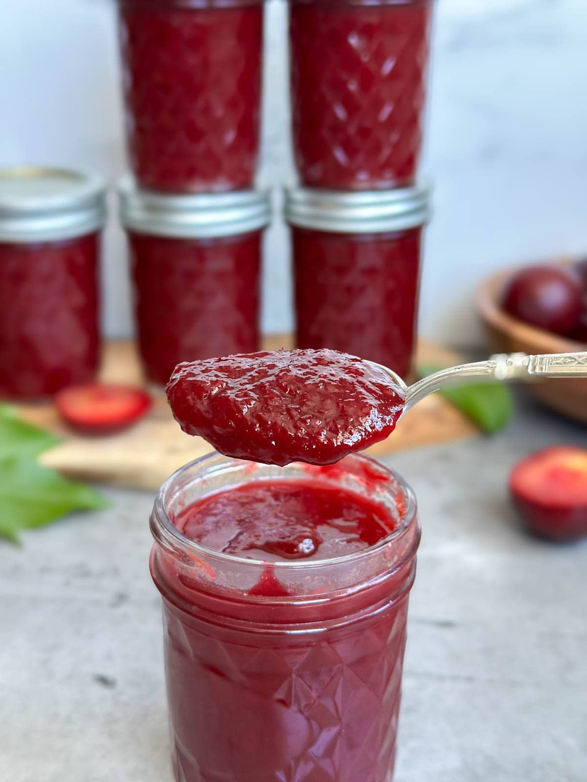 A large spoonful of plum jam is held above a half mason pint jar filled with the jam. Beyond are a number of jars of jam along with cut and whole fresh plums.