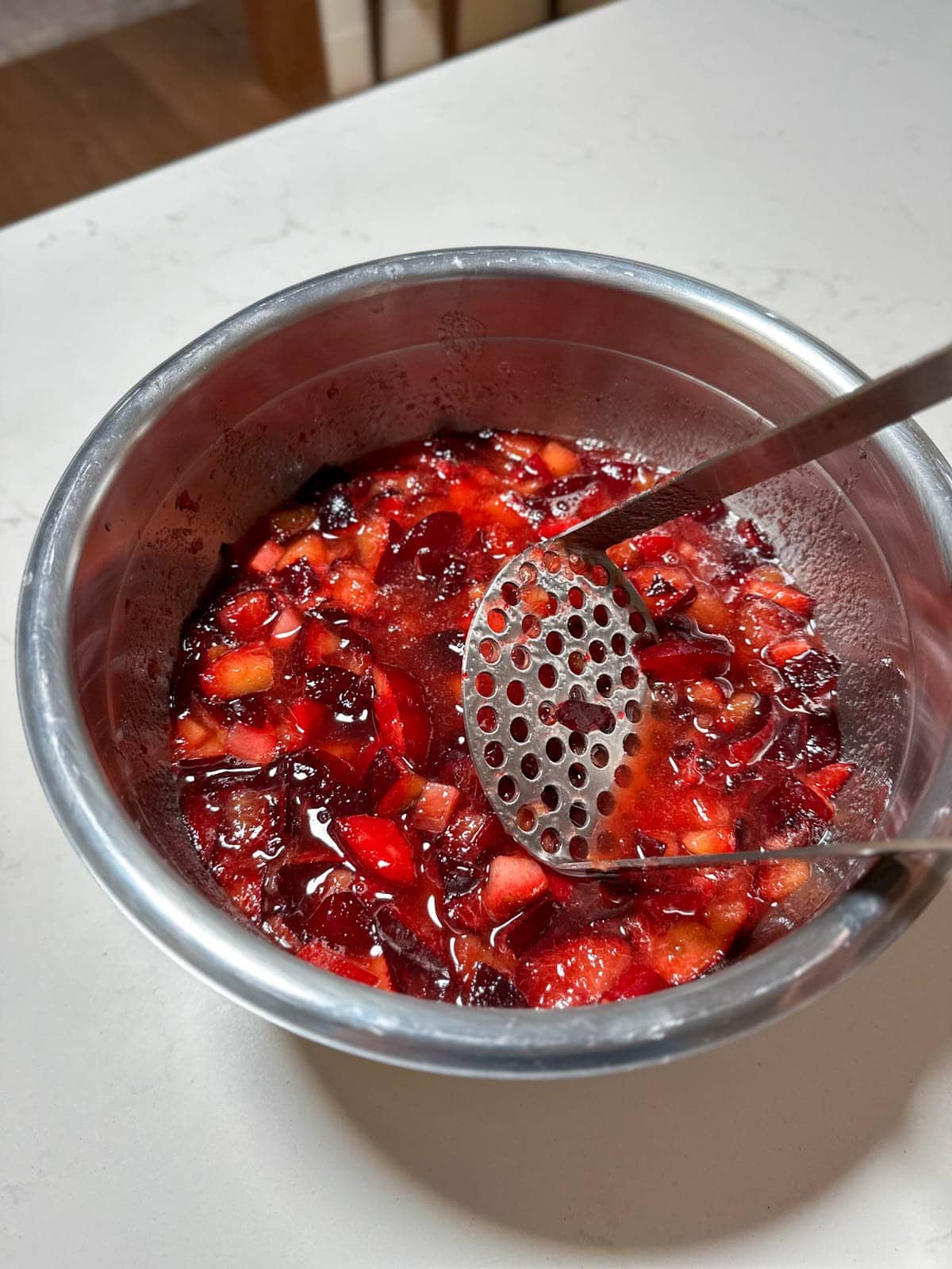 A stainless steel bowl full of fresh plums and sugar for the maceration step. A stainless steel potato masher is held just above the mixture.
