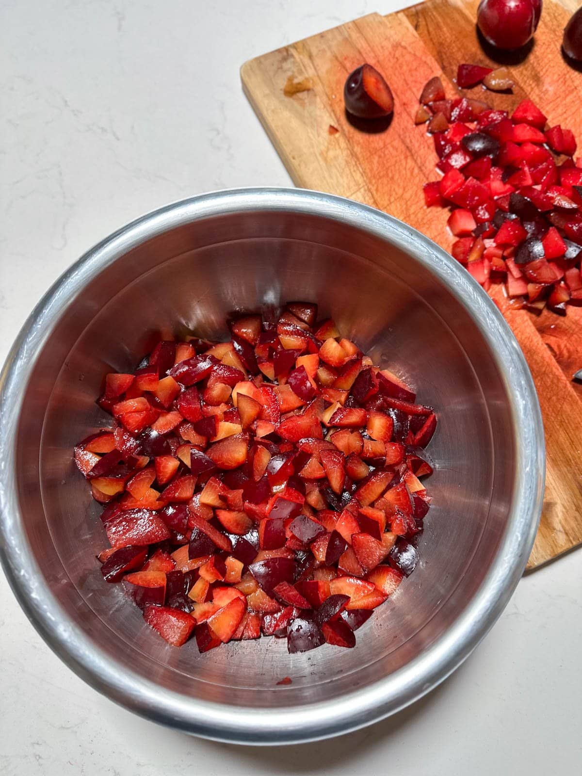 A stainless steel mixing bowl contains cut and processed fruit. A cutting board is next to the bowl with more processed fruit along with a few whole fruits.