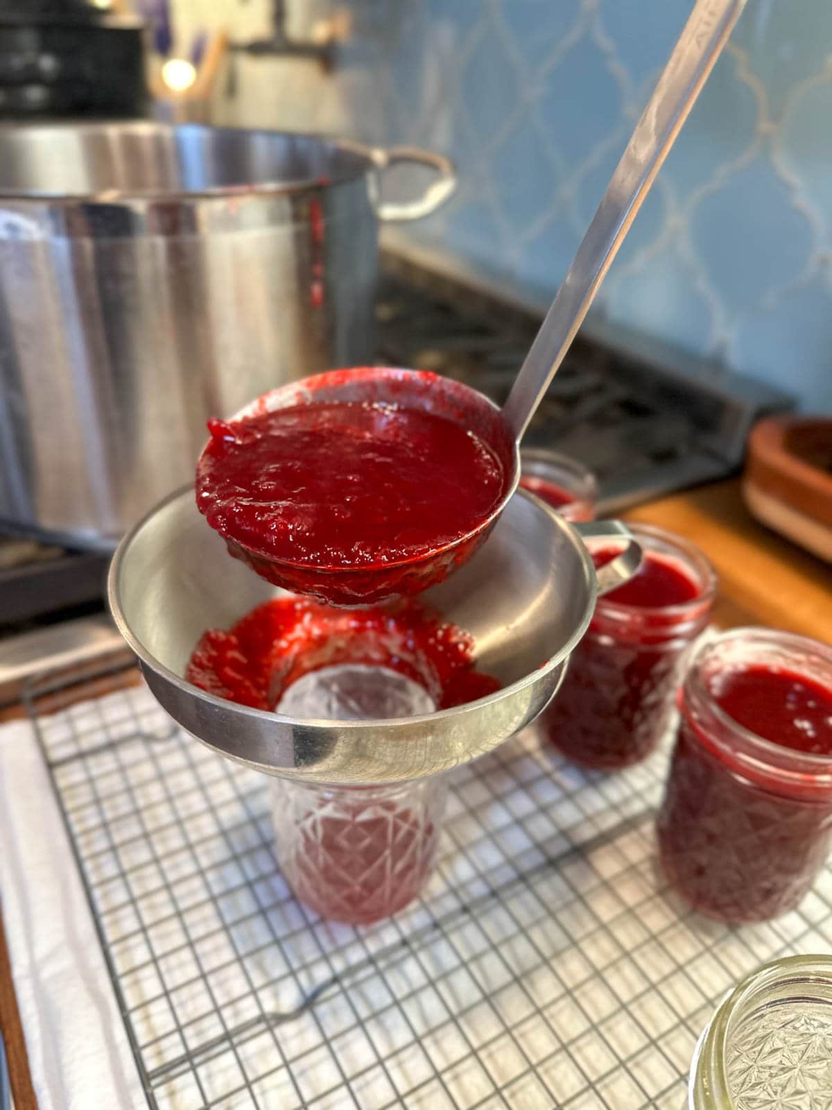 A ladle is spooning plum jam into half pint mason jars with a metal canning funnel on top of the jar. A few jars nearby are already full of plum jam.