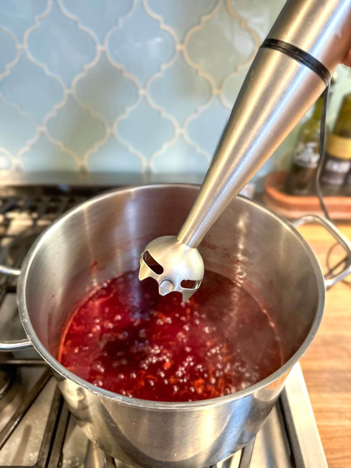 An immersion blender is held above a stock pot of plum jam cooking on a stove.