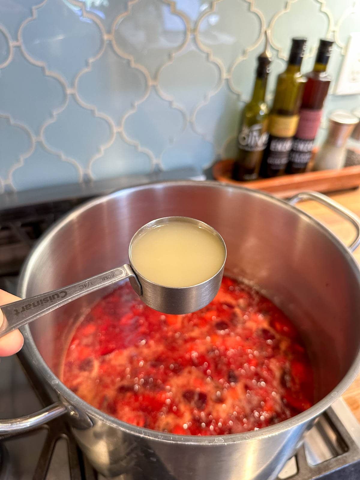 A 1/4 cup measurement of lemon juice is held above a stock pot of boiling fruit preserves.