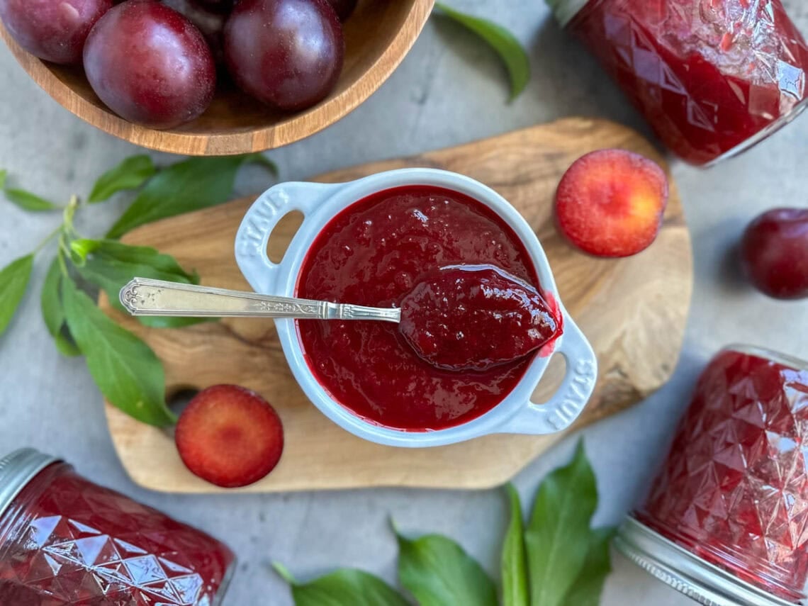 A small ramekin full of plum jam, a spoon is resting on top with a large dollop of jam inside of it. A few sliced plum, plum leaves, and a bowl of fresh plums garnish the surrounding area.