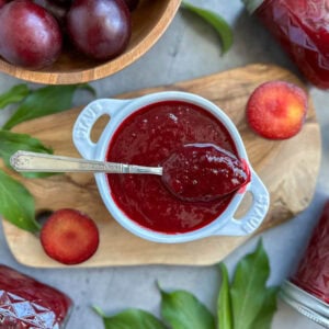 A small ramekin full of plum jam, a spoon is resting on top with a large dollop of jam inside of it. A few sliced plum, plum leaves, and a bowl of fresh plums garnish the surrounding area.