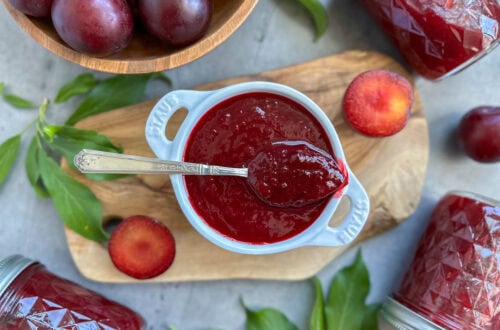 A small ramekin full of plum jam, a spoon is resting on top with a large dollop of jam inside of it. A few sliced plum, plum leaves, and a bowl of fresh plums garnish the surrounding area.
