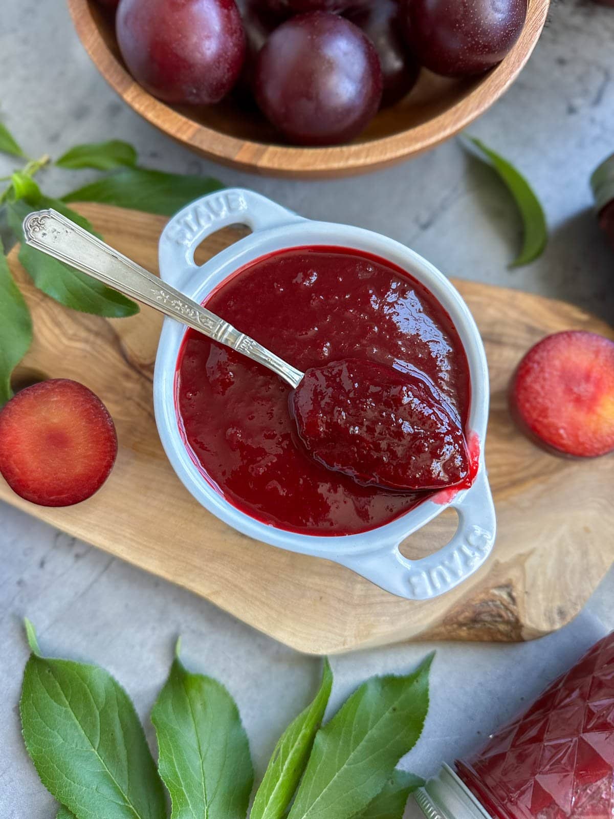 A small ramekin full of plum jam, a spoon is resting on top with a large dollop of jam inside of it. A few sliced plum, plum leaves, and a bowl of fresh plums garnish the surrounding area.