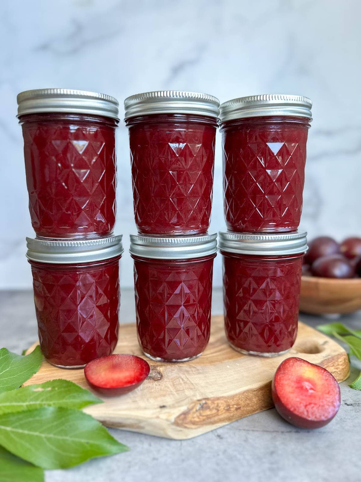 Six jars of fruit preserves sit in two rows of three, stacked on top of each other. A fruit that has been cut in half along with a few leaves from a tree surround the jars with a small bowl of fresh fruit in the background.