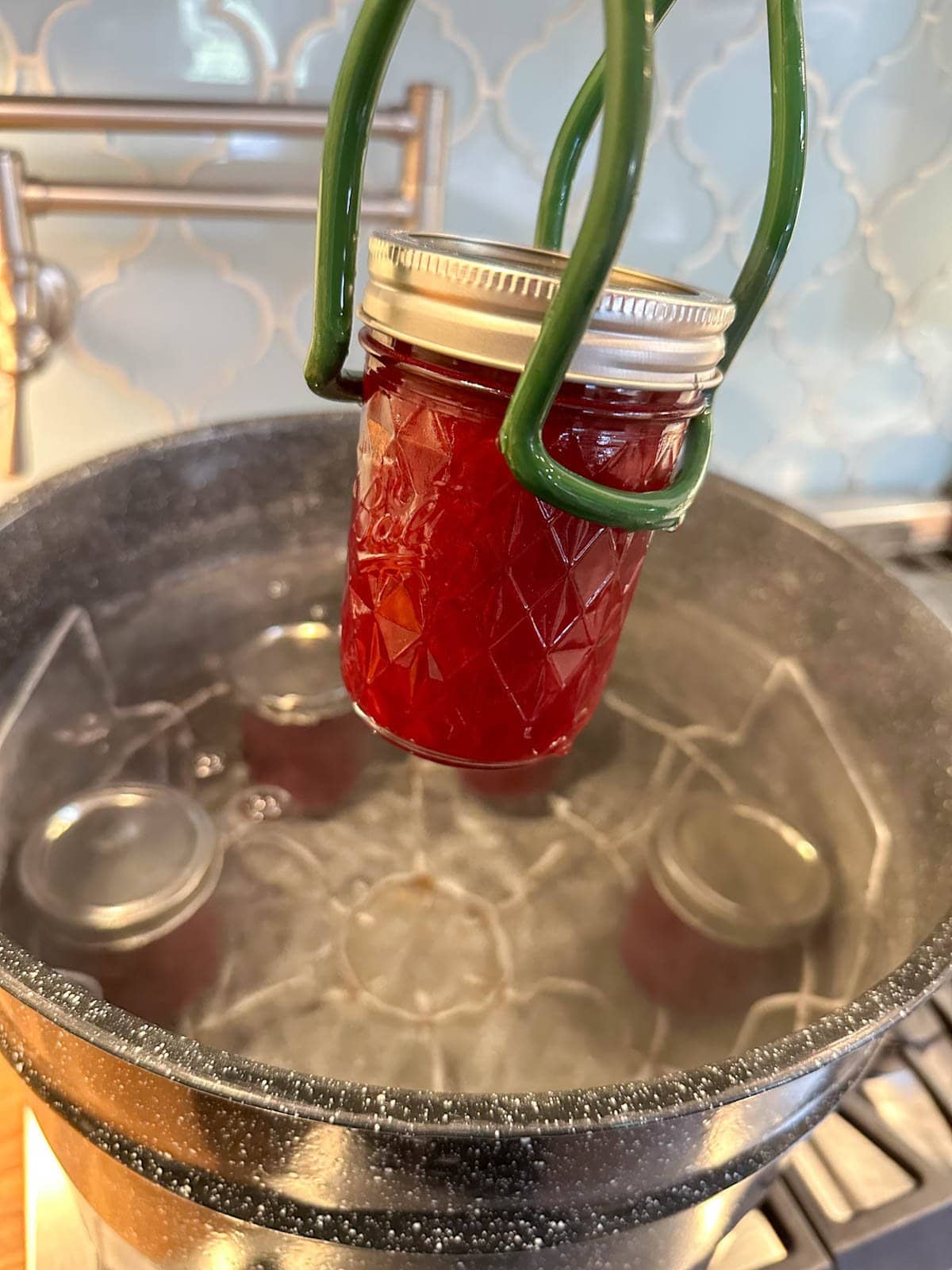 A jar lifter is holding a half pint mason jar of plum jam above a water bath canning pot with four jars of plum jam inside.