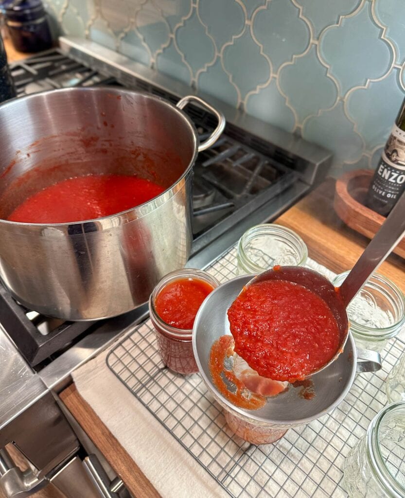 A stainless steel ladle ladling ketchup into pint jars with a canning funnel on top. A large stock pot full of finished ketchup is on the stove nearby.