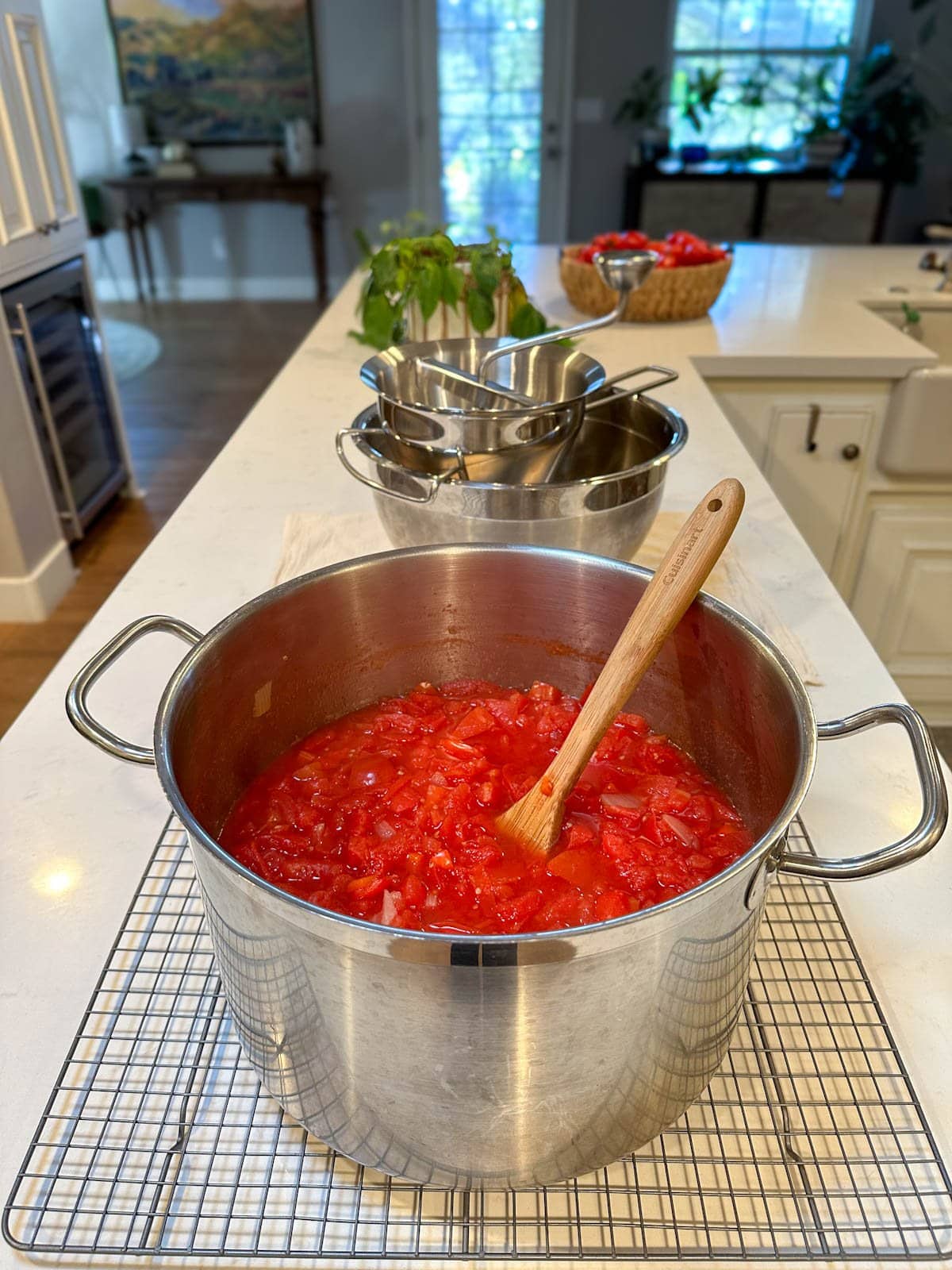 A stock pot full of cooked tomato, onion, and pepper. A food mill sits on top of a metal bowl beyond that will mill the ingredients to be made into homemade ketchup.