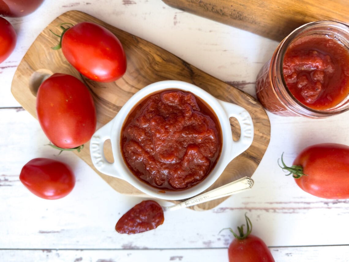 A small ramekin is full of homemade ketchup, a silver spoon is resting near the ramekin with a spoonful of ketchup inside. A few jars of finished ketchup surround the area along with a few fresh tomatoes.