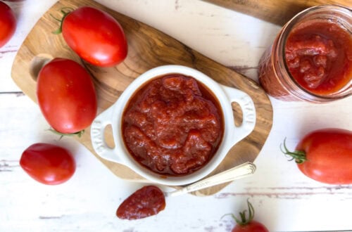 A small ramekin is full of homemade ketchup, a silver spoon is resting near the ramekin with a spoonful of ketchup inside. A few jars of finished ketchup surround the area along with a few fresh tomatoes.