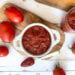 A small ramekin is full of homemade ketchup, a silver spoon is resting near the ramekin with a spoonful of ketchup inside. A few jars of finished ketchup surround the area along with a few fresh tomatoes.