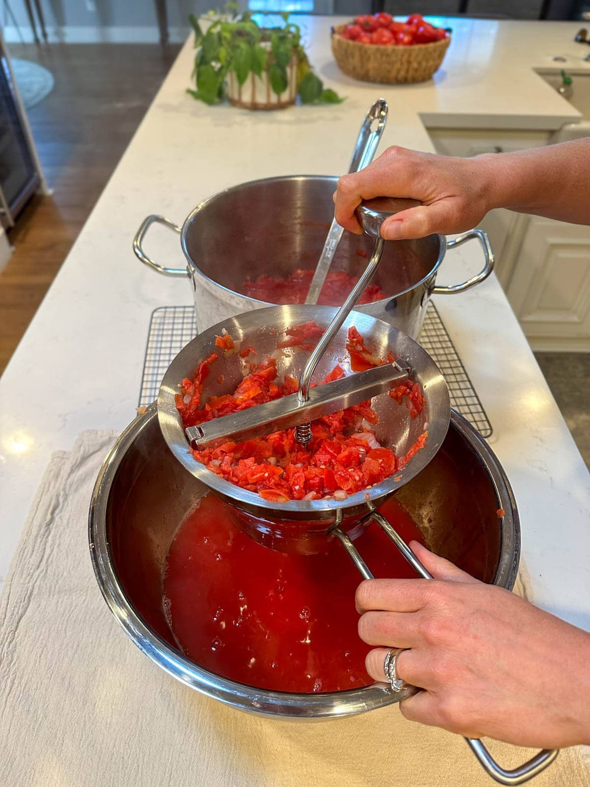 Tomato, pepper, and onions are being moved through the food mill with a large metal bowl below to catch the extracted juices. A large stock pot of the tomato, pepper, and onion mixture is right next to the mill.