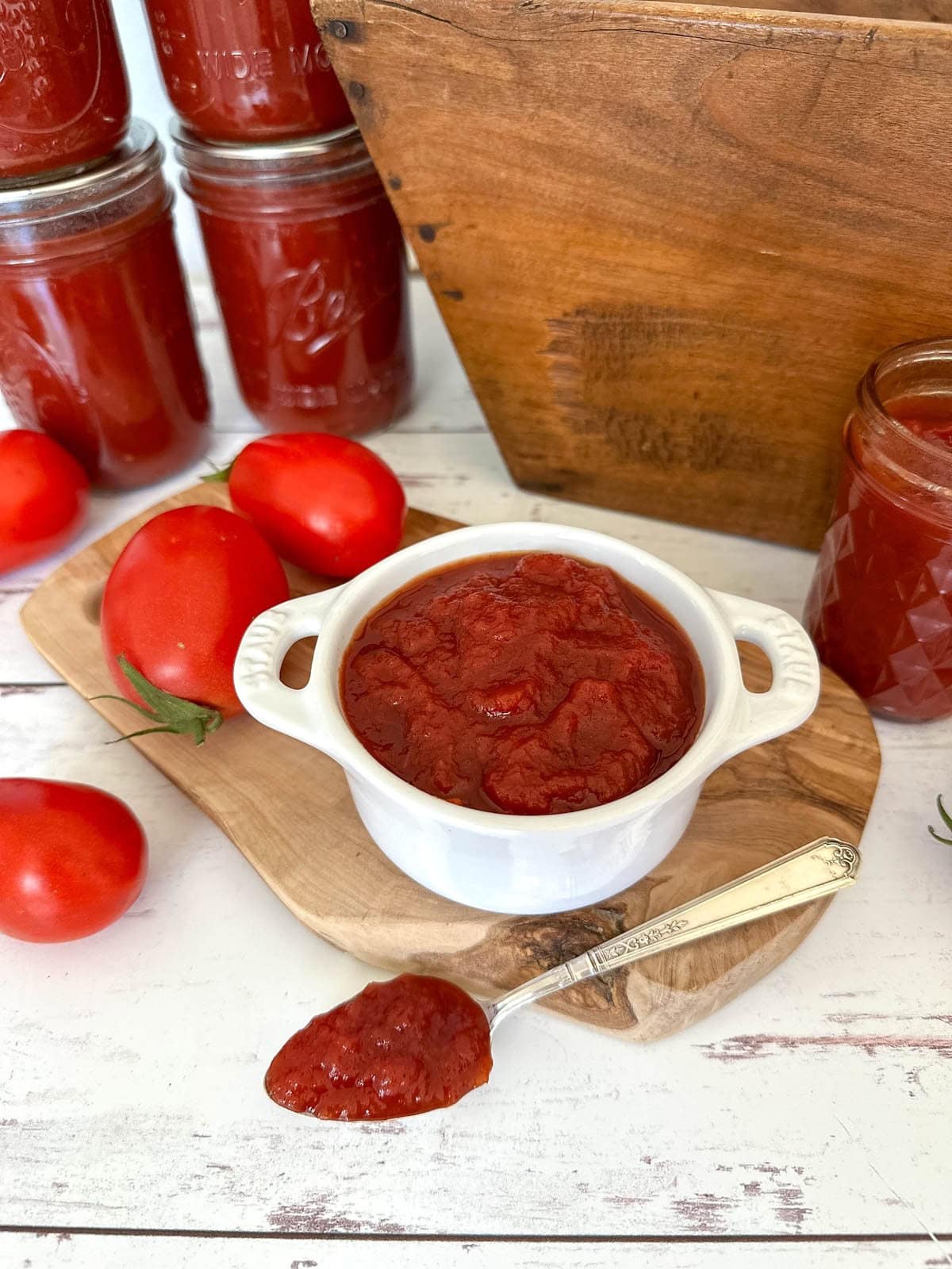 A small ramekin is full of homemade ketchup, a silver spoon is resting near the ramekin with a spoonful of ketchup inside. A few jars of finished ketchup surround the area along with a few fresh tomatoes.