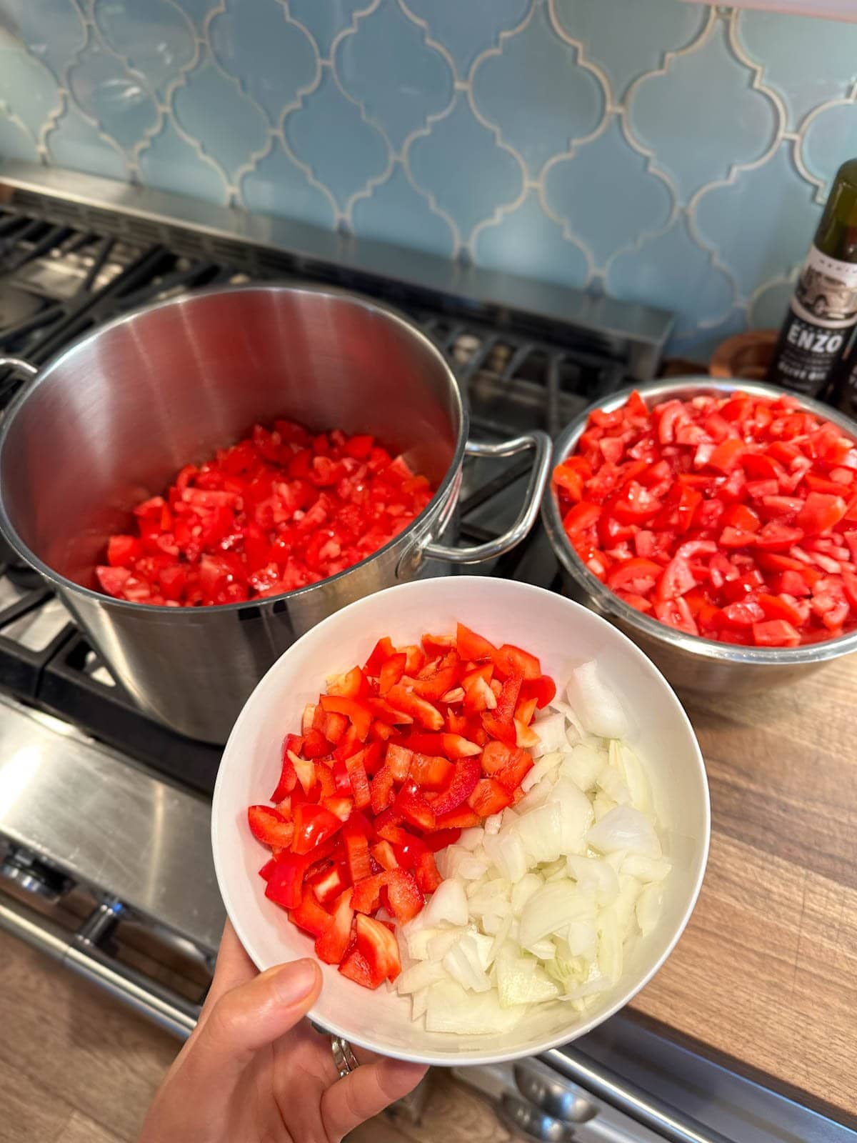 A small white bowl full of chopped onions and red bell pepper is held in front of a stock pot partially full of chopped tomatoes as well as a metal bowl heaped full of chopped tomatoes.