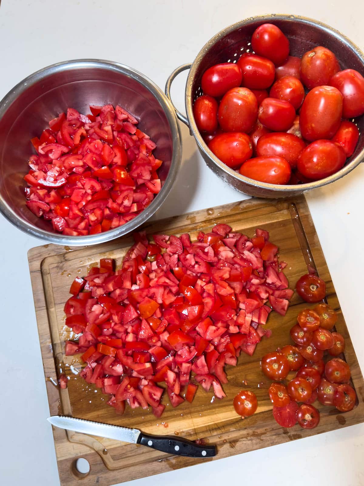 A large wood cutting board is covered in chopped tomatoes as well as the cores of the tomatoes. Two large bowls are above the cutting board, one is full of chopped tomatoes while the other contains fresh Roma tomatoes.