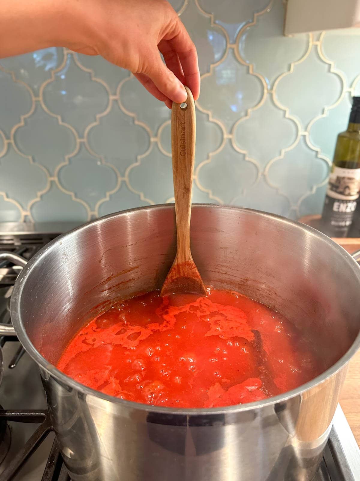 A large stainless steel stockpot full of tomato puree and other ingredients. A large wooden spoon is inserted vertically into the sauce so show how much is in the pot.