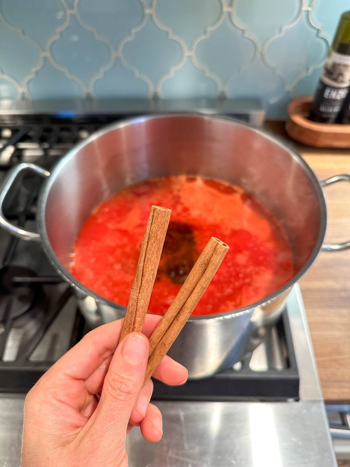 A hand holding two cinnamon sticks, a large pot full of ingredients simmering on the stove beyond.
