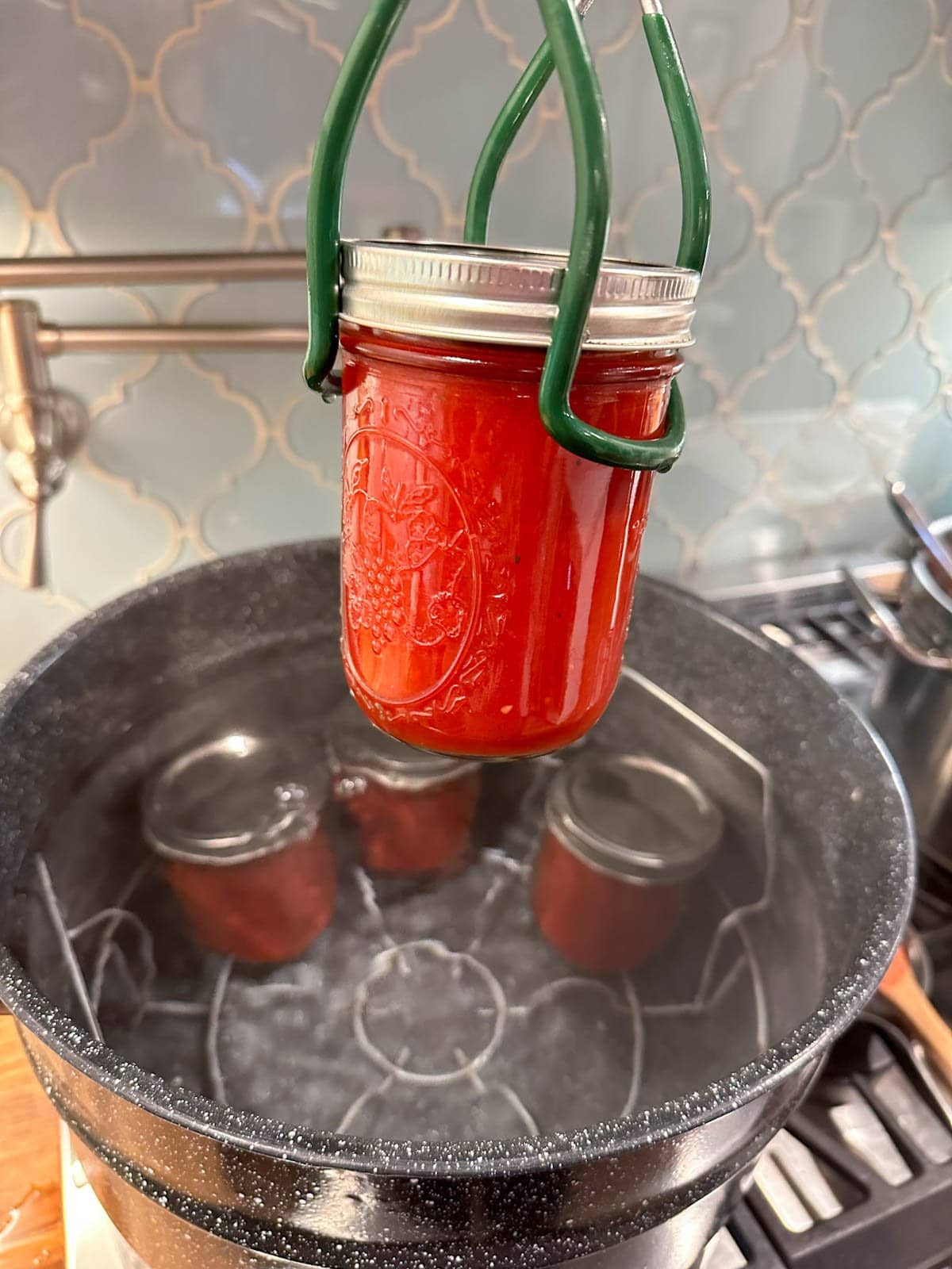 A jar lifter has a pint jar of homemade ketchup in its grasp, beyond is a large canning pot filed with boiling water and a few jars of ketchup.