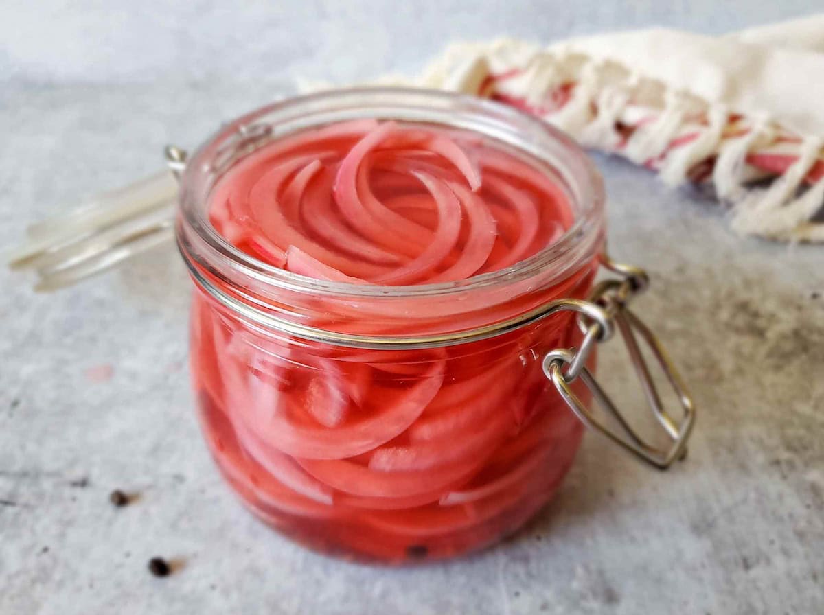 A small flip top jar full of pickled red onions, the onions appear pink in color along with the vinegar from the red onions.