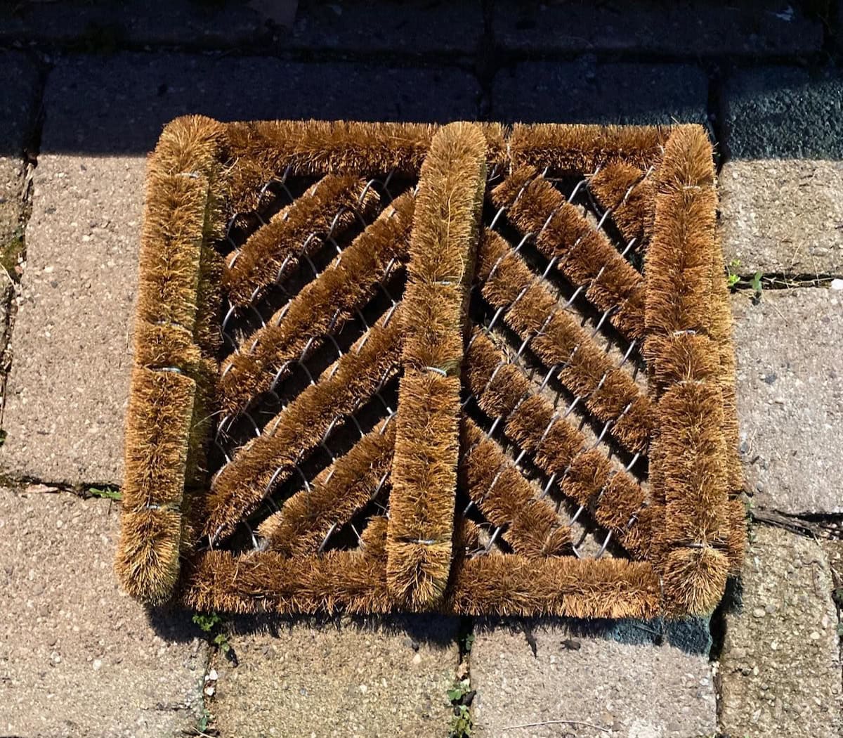 A boot scraper made of coconut fibers sits a top a brick hardscape. 
