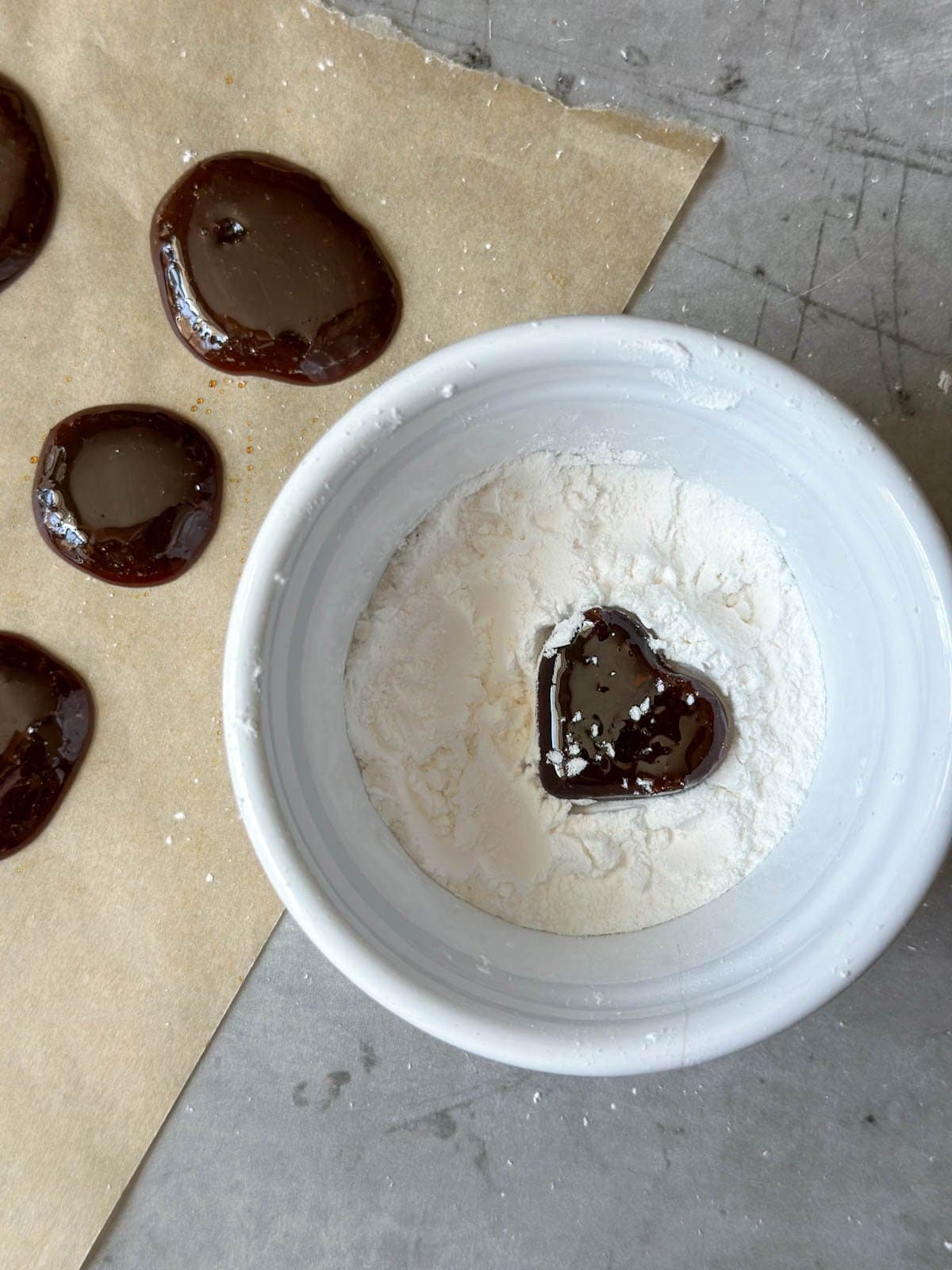 A homemade cough drop in the shape of a heart being coated in arrowroot powder inside of a small white ramekin.
