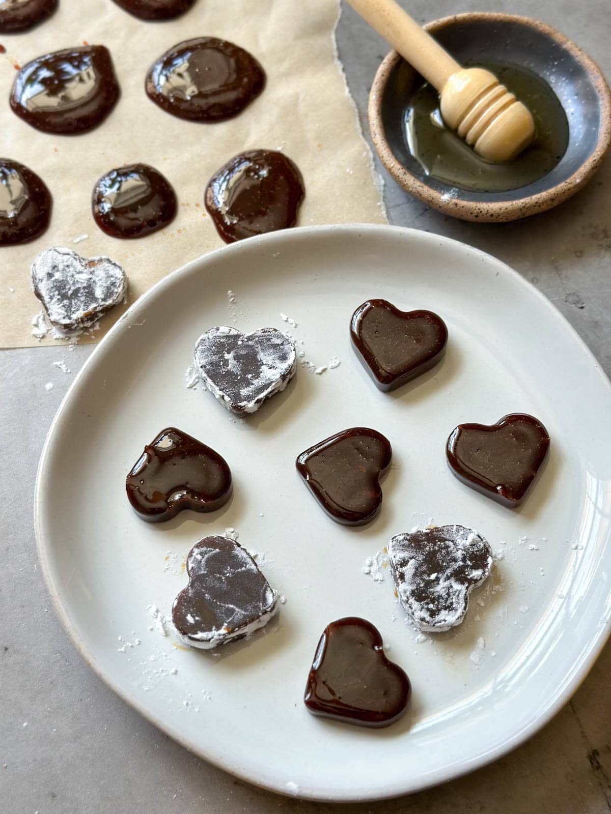 Heart shaped homemade cough drops are arranged on a ceramic plate while dollops are lined up on a piece of parchment paper. A small bowl has a small amount of honey in it with a wooden honey spoon inside of it.