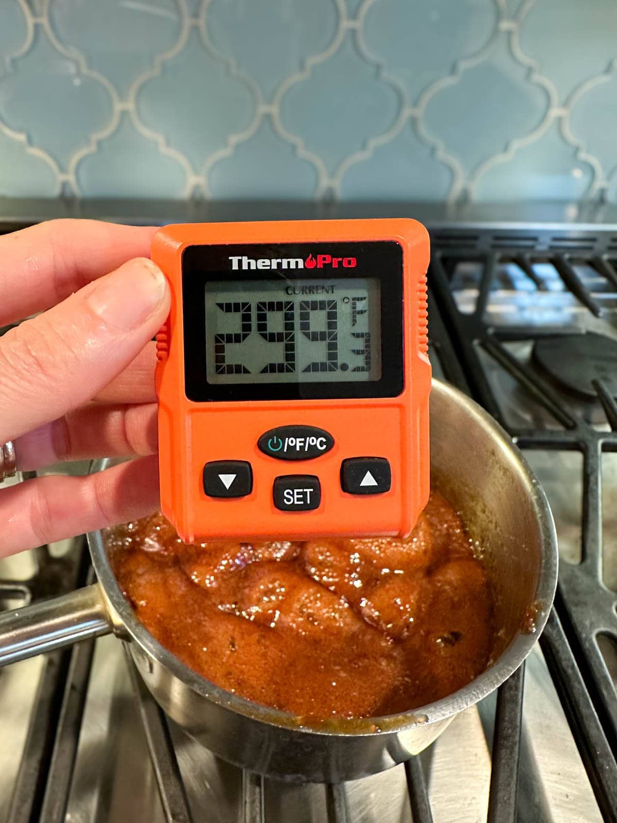 A candy thermometer reads 299 degrees F as a pot is on the stove with a bubbling brown mixture inside.