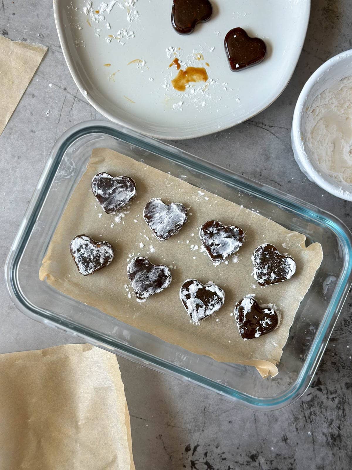 Homemade cough drops in the shape of hearts are lined on top of a piece of parchment paper inside of a glass container.