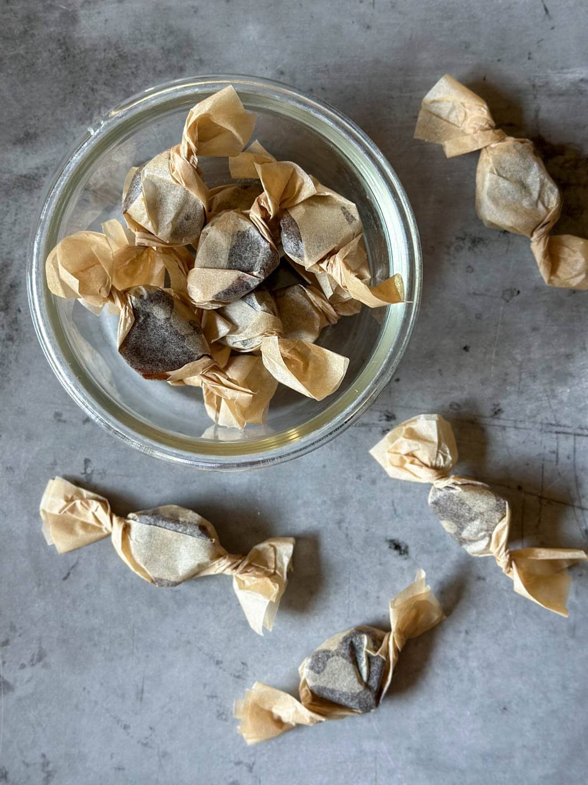 Many small homemade cough drops are wrapped in waxed paper similar to saltwater taffy, most are in a small glass bowl while a few are scattered around.