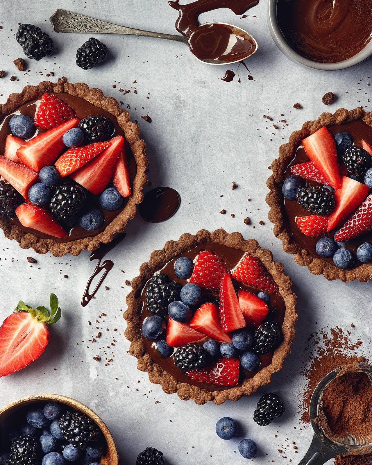Three small tarts topped with fresh berries are arranged in a triangle, fresh berries and cocoa powder garnish the surrounding area.