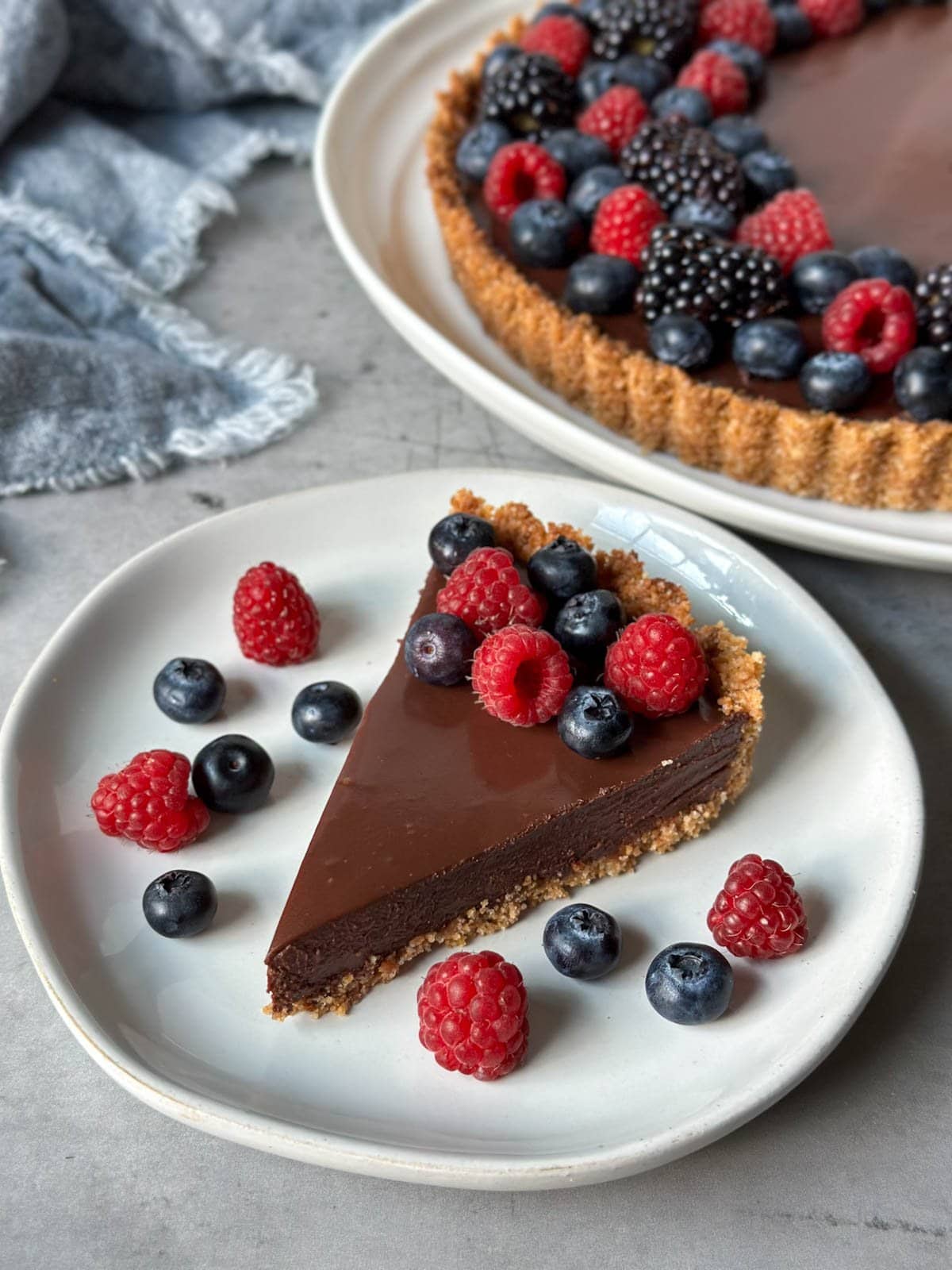 A slice of chocolate tart topped with fresh raspberries and blueberries is on a small plate. Just beyond is a large platter with the rest of the chocolate tart.