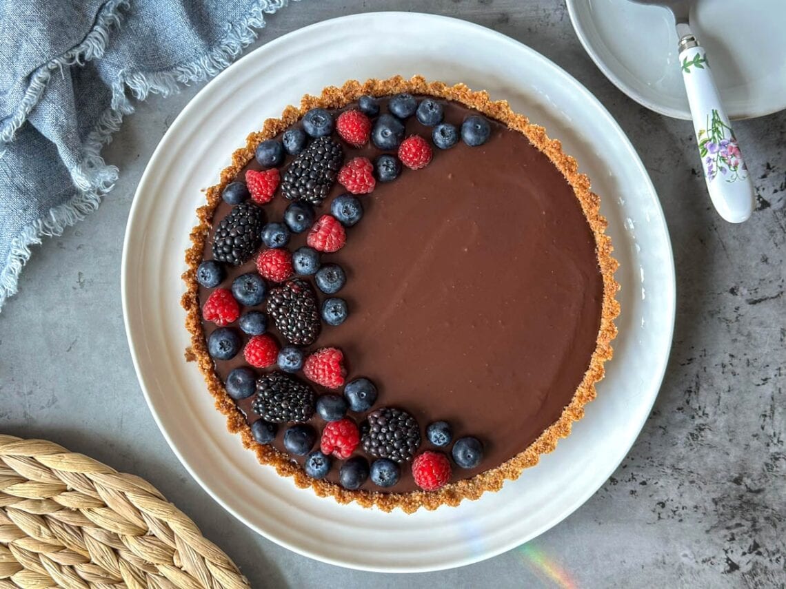 A dark chocolate tart with fresh berries arranged in a half crescent moon on top of it sit on a large ceramic serving plate.