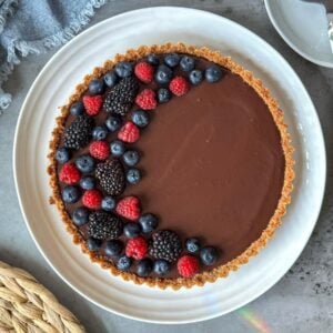 A dark chocolate tart with fresh berries arranged in a half crescent moon on top of it sit on a large ceramic serving plate.