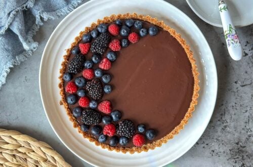 A dark chocolate tart with fresh berries arranged in a half crescent moon on top of it sit on a large ceramic serving plate.